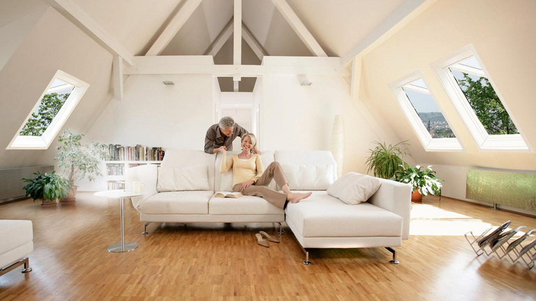 ein Paar wird in seiner Dachwohnung im Wohnzimmer gezeigt, die Dachfenster an den Seiten sind geöffnet zum Lüften